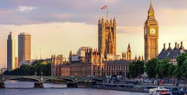 Das Bild zeigt die Themse mit dem Palace of Westminster und dem berühmten Uhrturm Big Ben im Hintergrund. Die Gebäude sind von warmem Licht erleuchtet, während der Himmel in sanften Farben leuchtet. Ein paar Boote sind auf dem Wasser zu sehen.