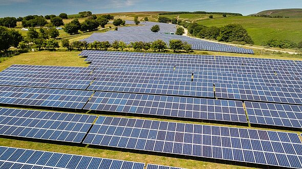 Aerial view of a large solar farm, featuring numerous rows of solar panels arranged neatly across a green field. The panels reflect sunlight, indicating energy production, while trees line the perimeter, with hills visible in the background.