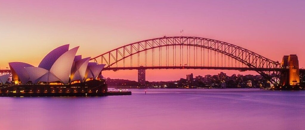 Das Bild zeigt das Sydney Opera House mit seinen charakteristischen, segelartige Dächern und die Sydney Harbour Bridge, die über den ruhig glühenden Hafen spannt. Der Himmel hat einen sanften, lila Farbton während der Dämmerung.