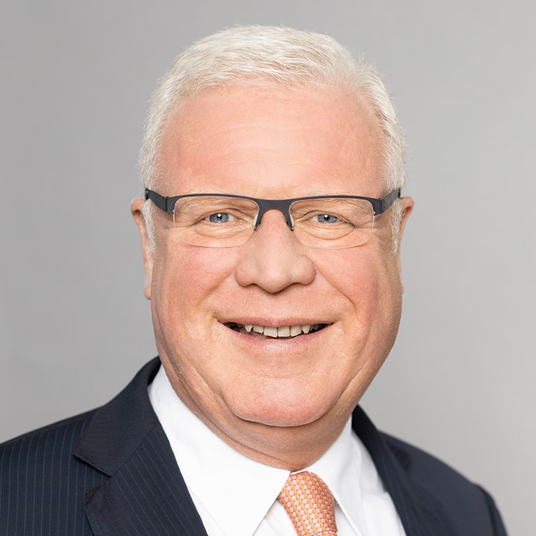 A smiling middle-aged man with short, light hair and glasses is dressed in a dark suit with a white shirt and an orange tie. He is facing the camera against a light gray background, projecting a friendly and professional demeanor.