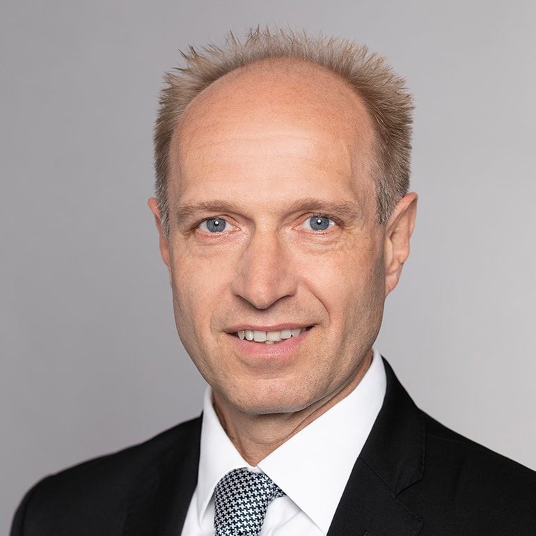 A professional headshot of a man with short, light brown hair and blue eyes. He is wearing a dark suit and a white shirt with a patterned tie. He smiles confidently against a light gray background.