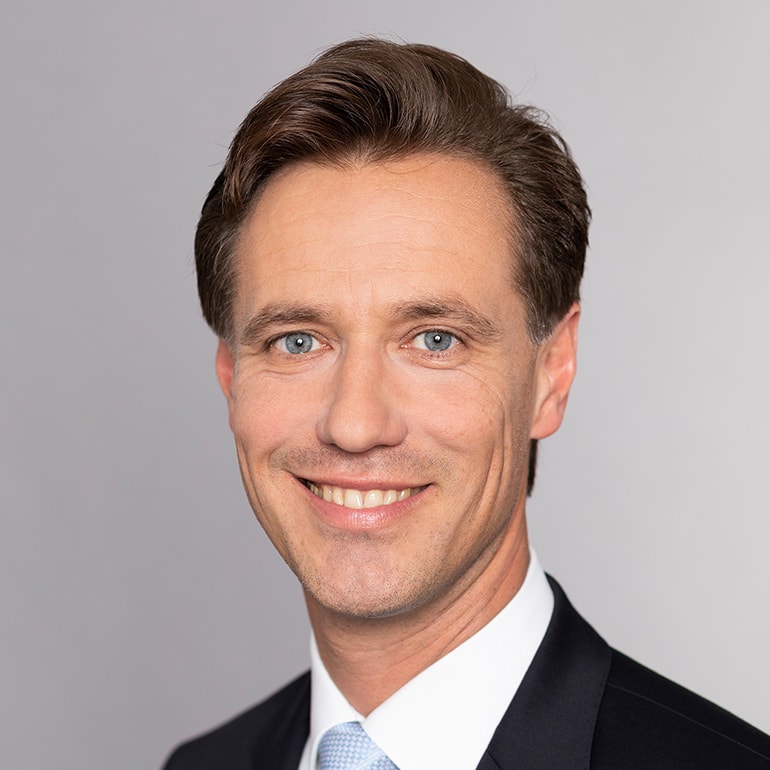 A man with short, styled brown hair and blue eyes smiles directly at the camera. He is dressed in a suit and tie, conveying a professional appearance. The background is a light gray, focusing attention on his friendly expression.