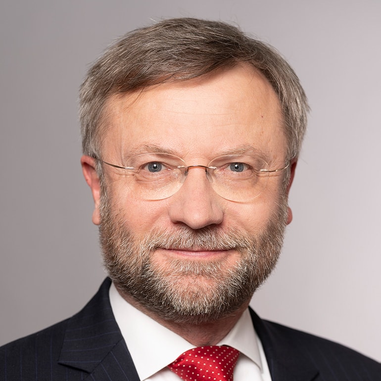 A middle-aged man with a beard and glasses smiles at the camera. He is wearing a suit with a white shirt and a red tie. The background is a plain light color, highlighting his professional appearance.