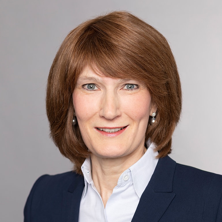 A woman with shoulder-length, brown hair smiles warmly at the camera. She wears a dark blazer over a light shirt. The background is a soft gray, which highlights her professional appearance.