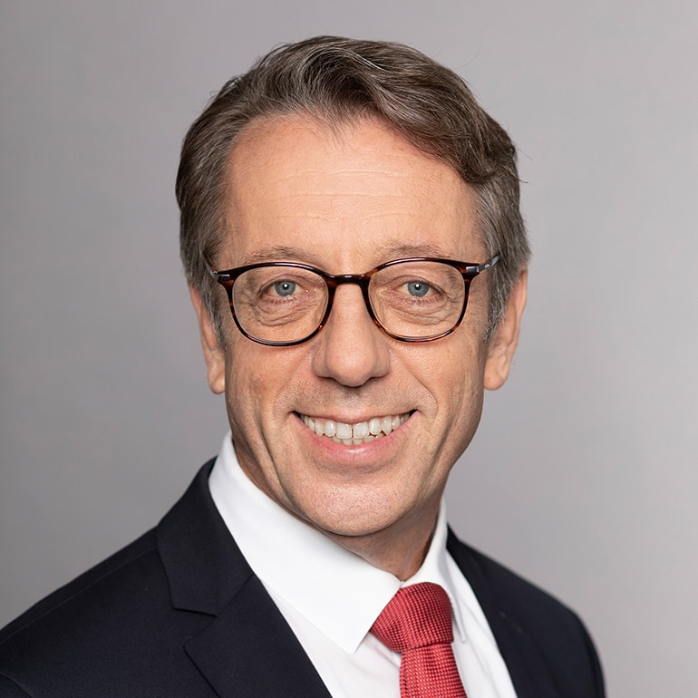 A professional man with short, light brown hair smiles warmly at the camera. He is wearing glasses and a dark suit with a white shirt, complemented by a red tie. The background is a neutral gray, highlighting his facial features.