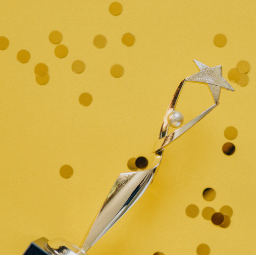 A shiny gold trophy features a swirling design topped with a star and a pearl, set against a bright yellow background. Small confetti pieces in gold are scattered around, enhancing the celebratory theme of achievement and recognition.