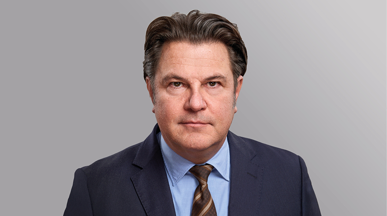 A middle-aged man with dark hair and a serious expression is wearing a suit and tie. His shirt is light blue, and he has a neutral background, making him the focal point of the image.