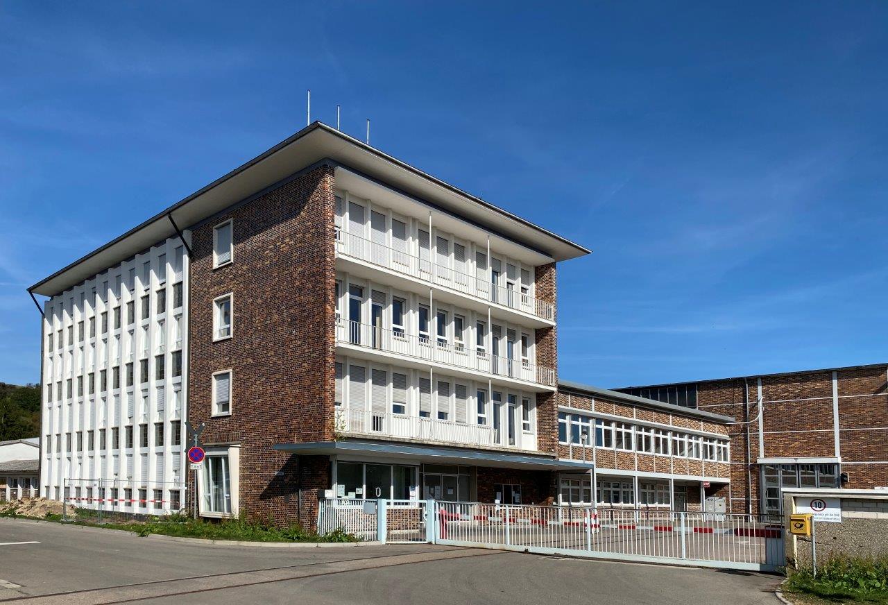 A modern, multi-story building with a mix of red brick and white facade, featuring large windows and balconies. The entrance is marked by a glass door and a gated area, set against a clear blue sky.