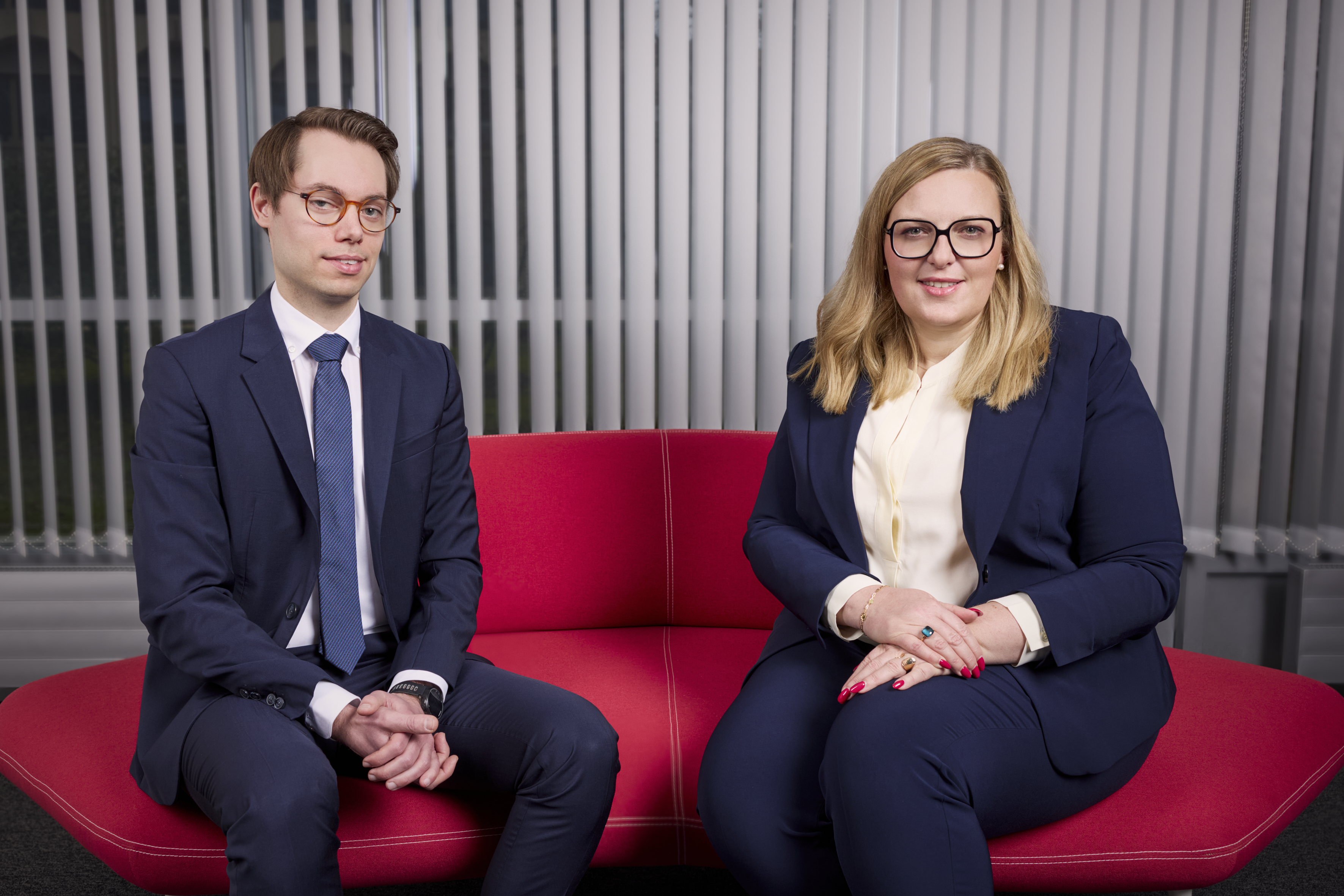 A man and a woman sit side by side on a red couch. Both are dressed in formal business attire, with the woman wearing glasses and having medium-length blonde hair. They are positioned against a backdrop of vertical blinds, exuding a professional atmosphere.