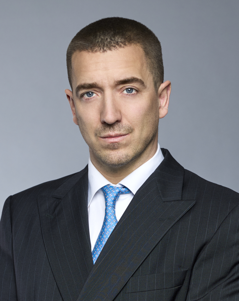 Porträt eines Mannes in einem gestreiften Anzug mit einer blauen Krawatte. Er hat kurzes, braunes Haar und sieht direkt in die Kamera. Der Hintergrund ist einfarbig und unauffällig.