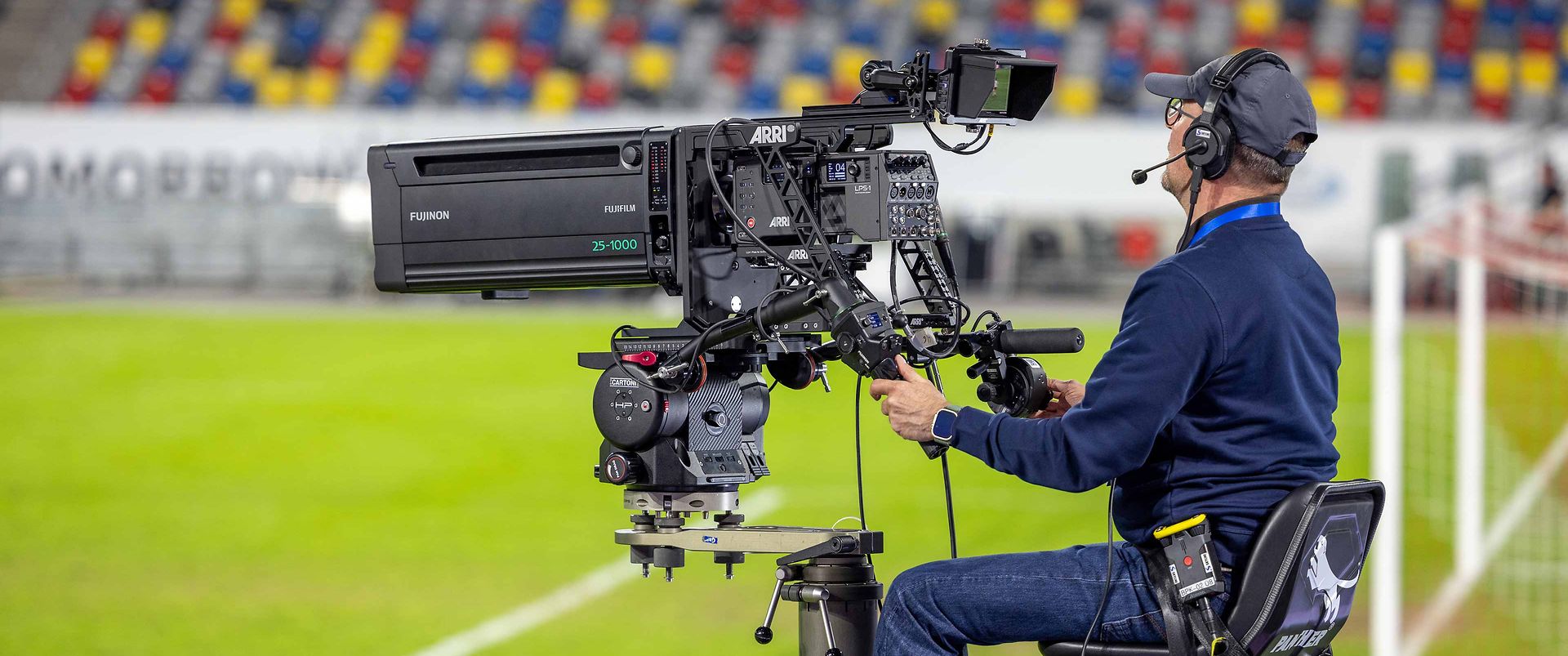 Ein Kameramann sitzt auf einem Stuhl und bedient eine professionelle Fernseh-Kamera auf einem Sportplatz. Die Kamera ist auf einem stabilen Stativ montiert. Im Hintergrund sind farbige Sitze im Stadion zu sehen. Der Mann trägt Kopfhörer und hat einen konzentrierten Gesichtsausdruck.