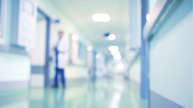 A blurred hospital corridor with soft lighting. A healthcare professional in a white coat stands by a door, suggesting an environment focused on medical care. The atmosphere conveys a sense of clinical activity and support.