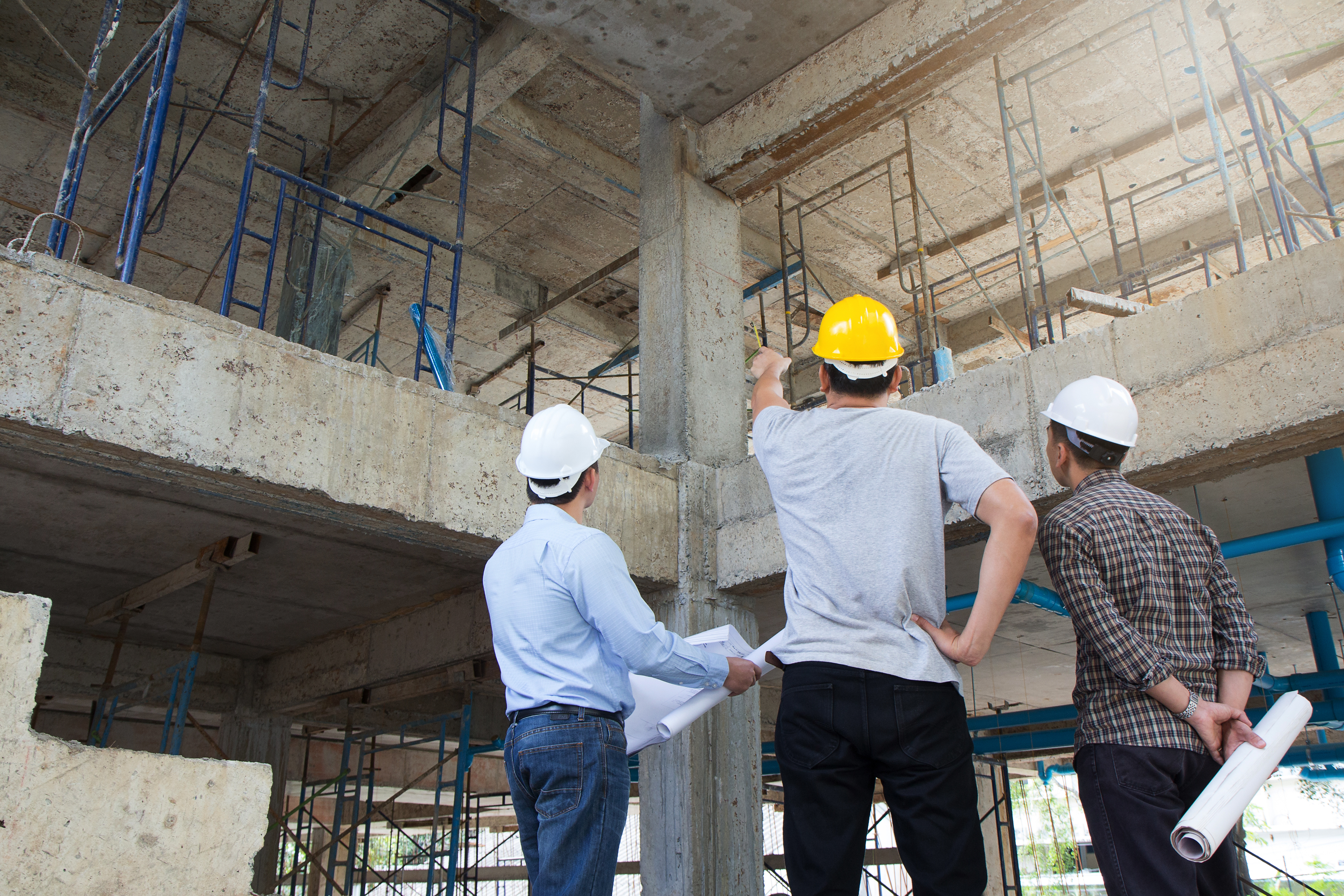 Three construction workers, wearing white helmets and one in a yellow helmet, examine building plans while discussing a concrete structure under construction. Scaffolding and construction elements are visible in the background, indicating an active construction site.