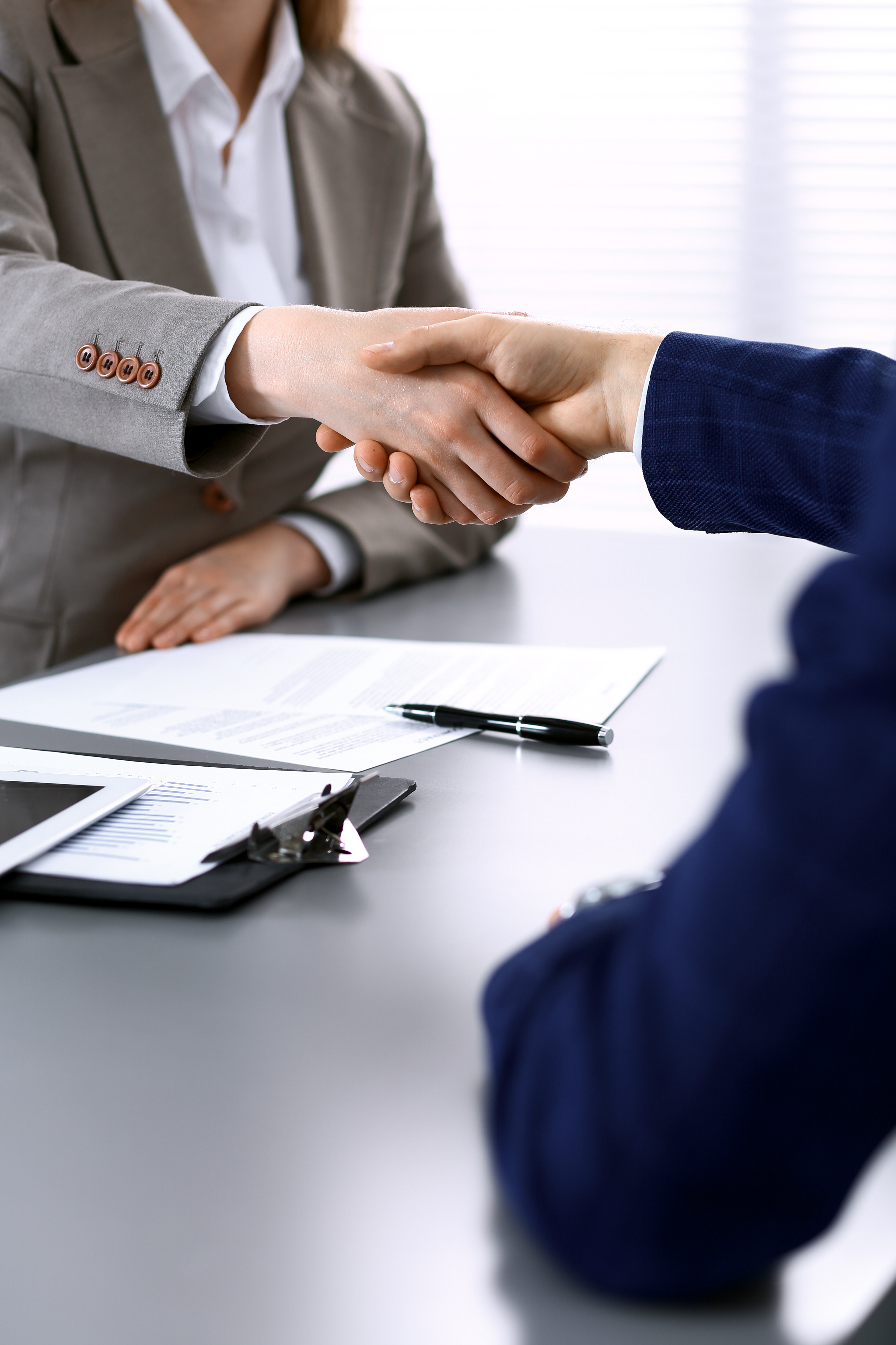A business meeting is taking place. Two people are shaking hands across a table, signaling an agreement or collaboration. Documents and a tablet with graphs are visible on the table, indicating a professional setting.
