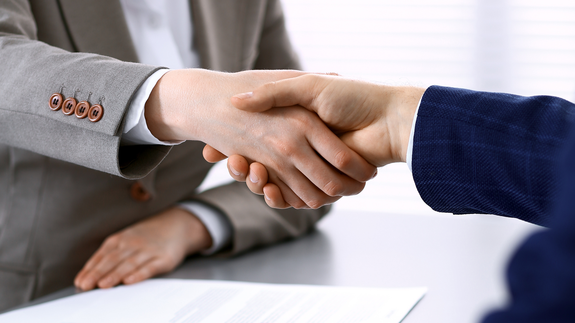A close-up of two hands engaged in a handshake, symbolizing agreement or partnership. One hand is in a gray suit, while the other is in a blue suit. The background is softly blurred, suggesting a professional setting.