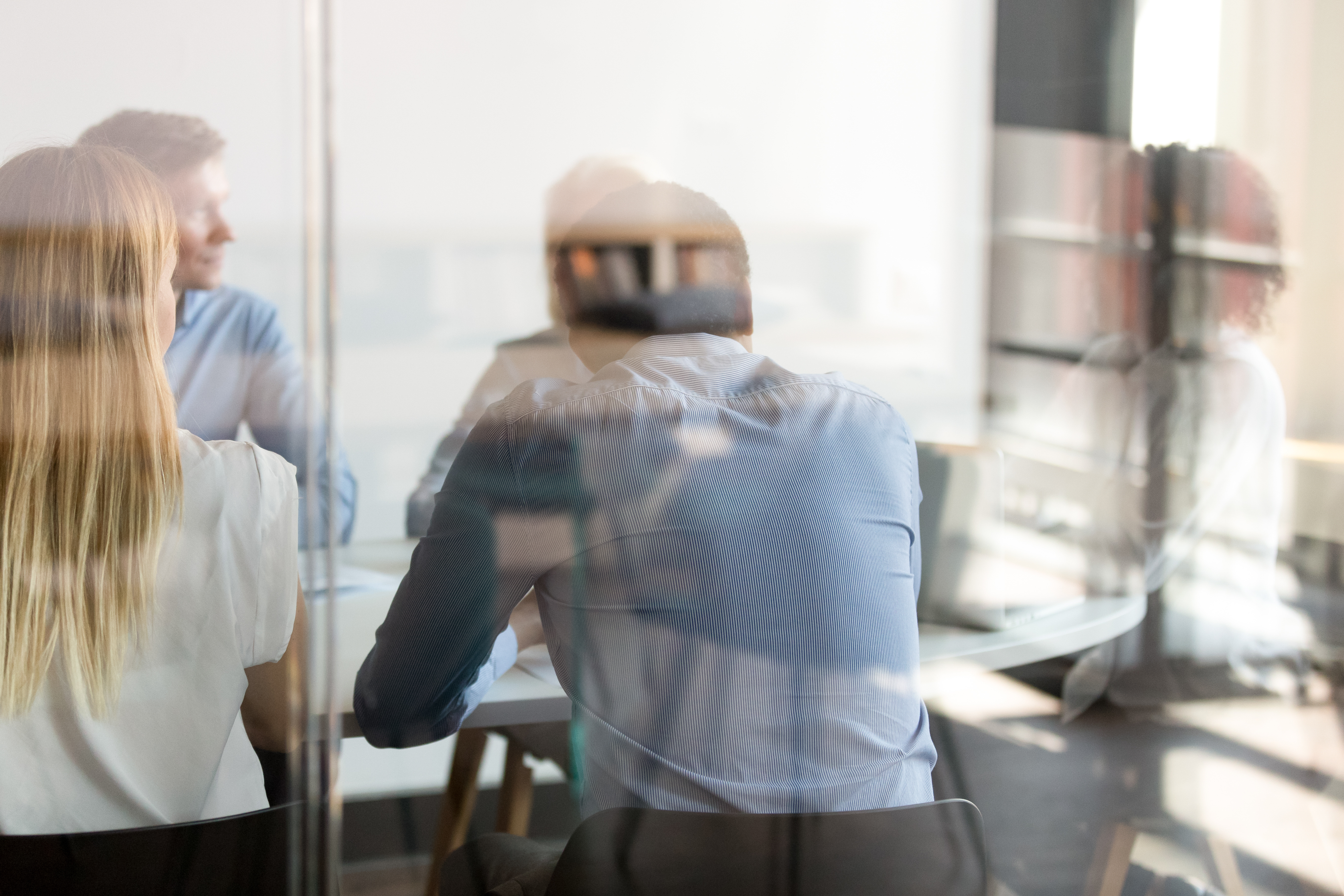 A group of professionals in a meeting room is depicted, with four individuals visible from the back. They are engaged in discussion, facing a table with documents. The environment is modern and bright, creating a collaborative atmosphere.