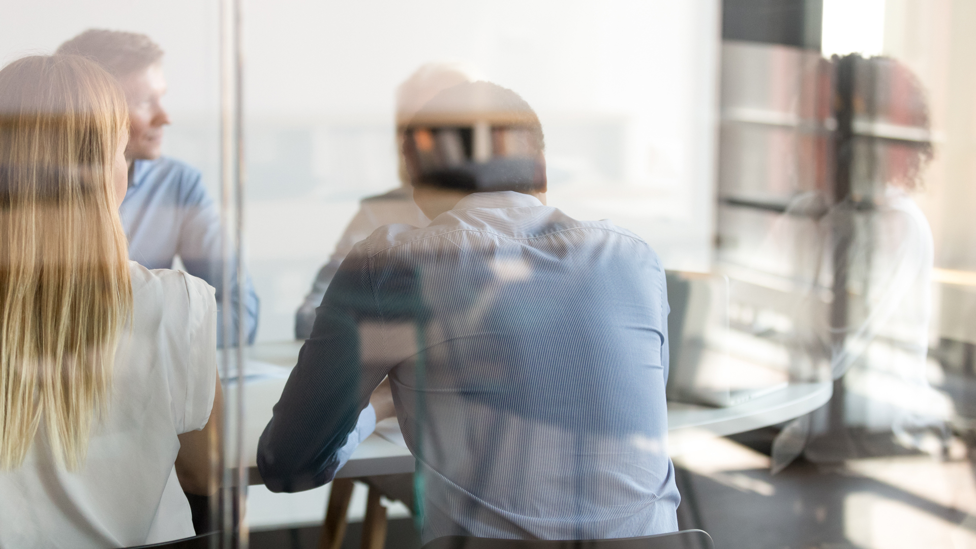A group of people is seated around a conference table, engaged in discussion. The focus is on the back of individuals, highlighting a collaborative meeting environment. Reflections from glass create a layered visual effect, enhancing the sense of a modern workspace.