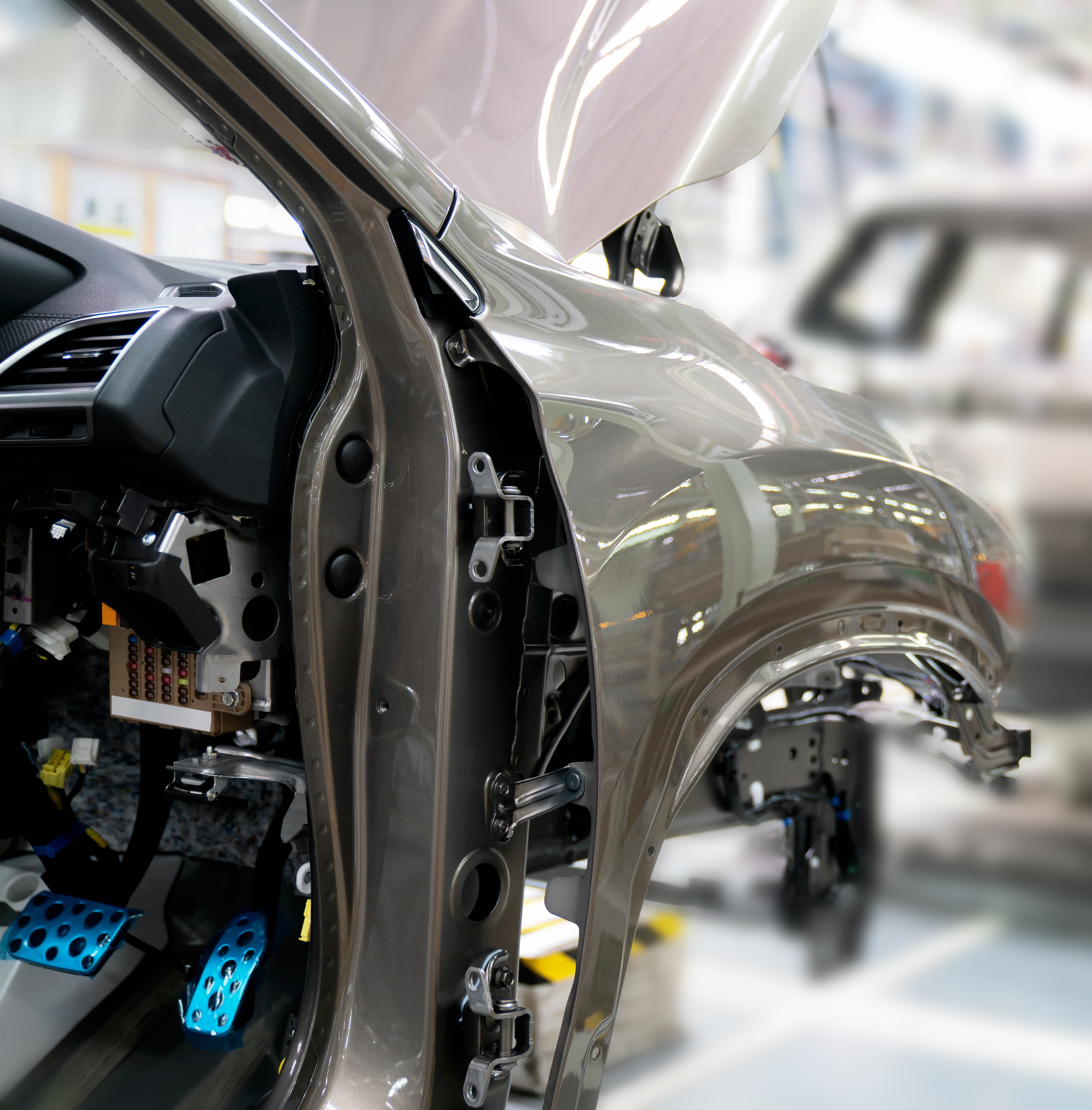 An image of a car's partial body frame in a factory setting, focusing on the open door area. The interior reveals components like pedals and wiring, showcasing the assembly process without fully illustrating the vehicle. The background is blurred, emphasizing the frame details.