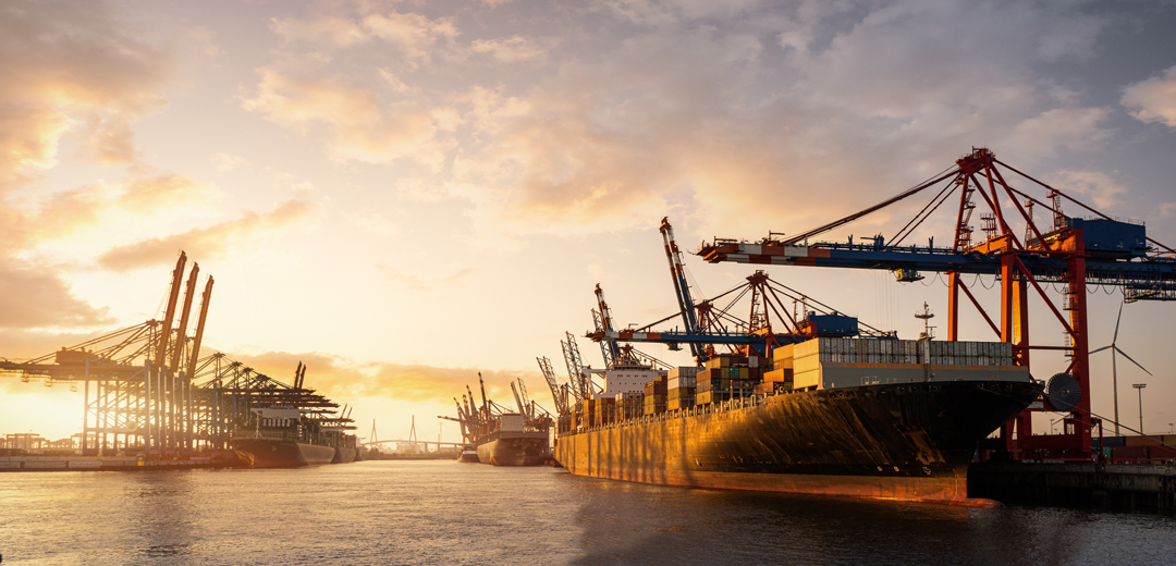The image shows a busy port at sunset, featuring large cargo ships docked alongside cranes. The sky is filled with warm colors as the sun sets, illuminating the scene, with several cranes positioned above the ships for loading and unloading cargo.