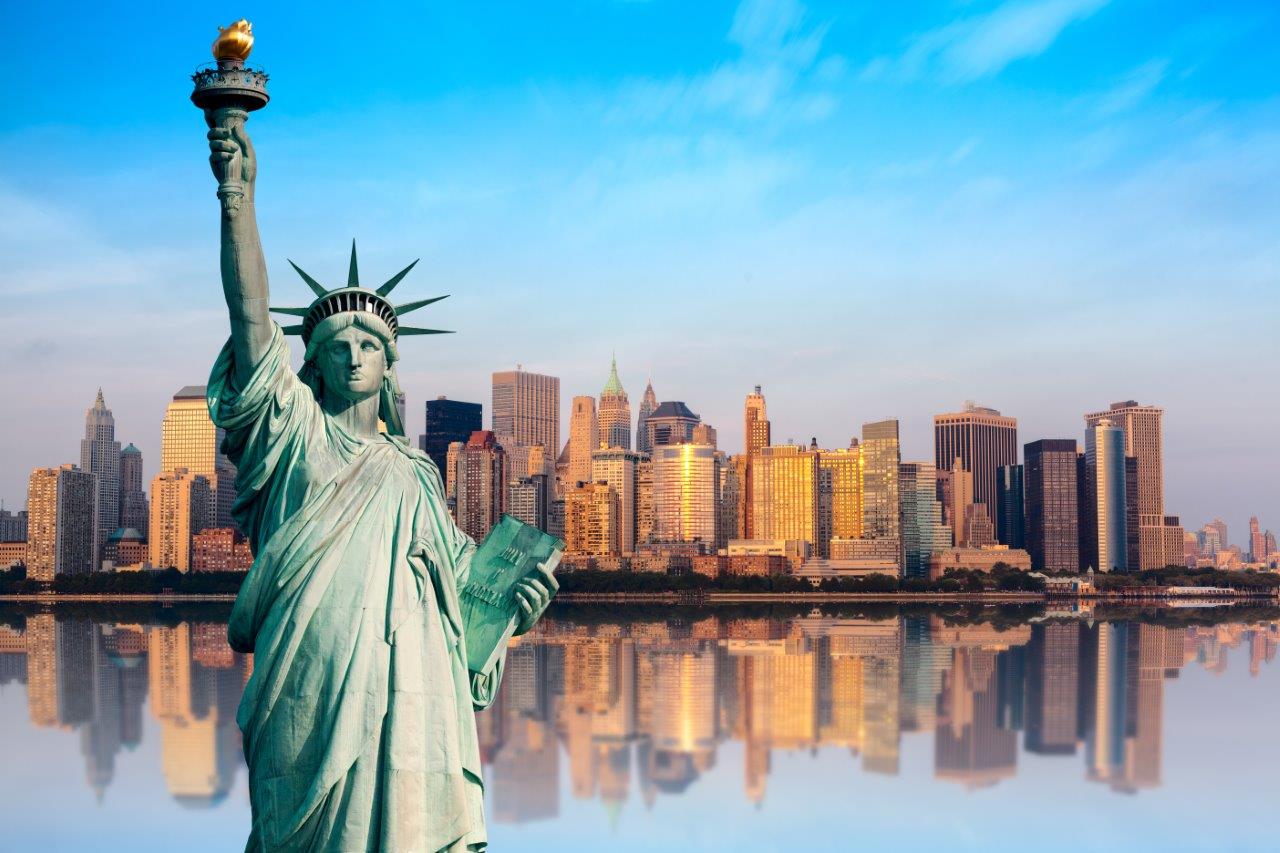 Die Statue der Freiheit, ein bedeutendes Symbol für Freiheit und Hoffnung, steht im Vordergrund. Im Hintergrund ist die Skyline von New York City zu sehen, die von hohen Gebäuden geprägt ist. Der Himmel ist blau mit einigen Wolken.