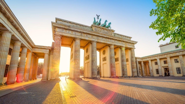 Das Bild zeigt das Brandenburger Tor in Berlin bei Sonnenuntergang. Die monumentale Struktur aus Säulen und Skulpturen ist erleuchtet von warmem Licht, während die Umgebung ruhig und einladend wirkt.
