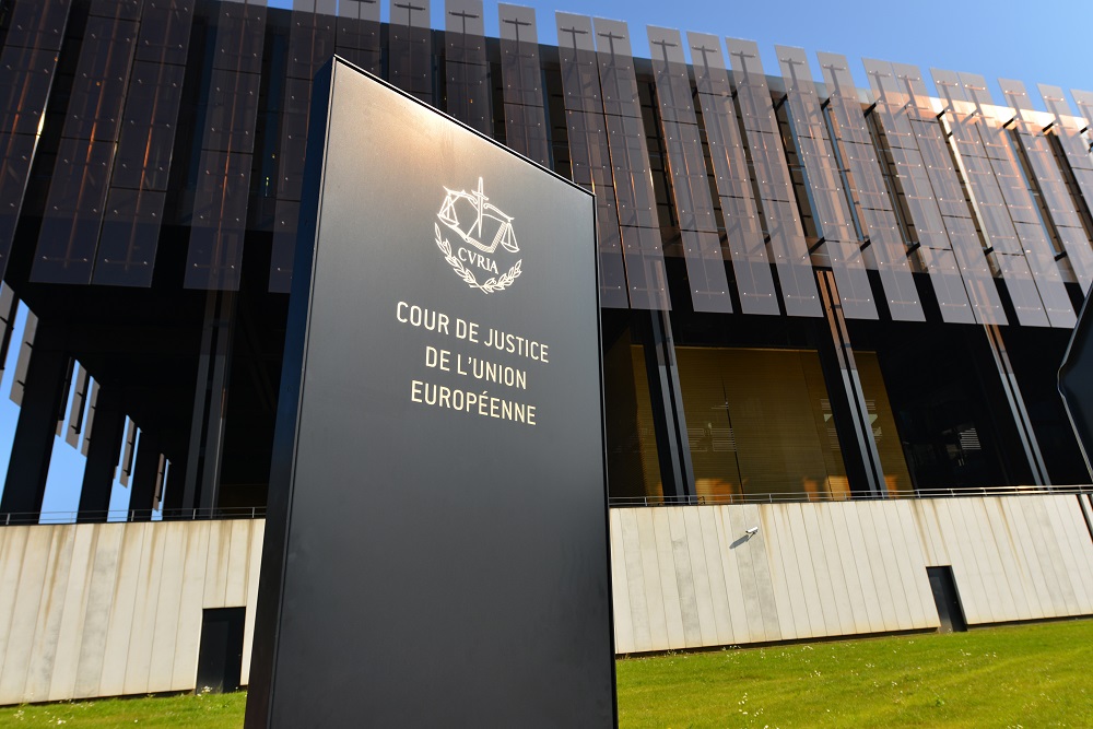 A black sign with golden text reads "COUR DE JUSTICE DE L'UNION EUROPÉENNE" next to the modern glass building of the Court of Justice of the European Union. The structure features large glass windows and a contemporary architectural design.