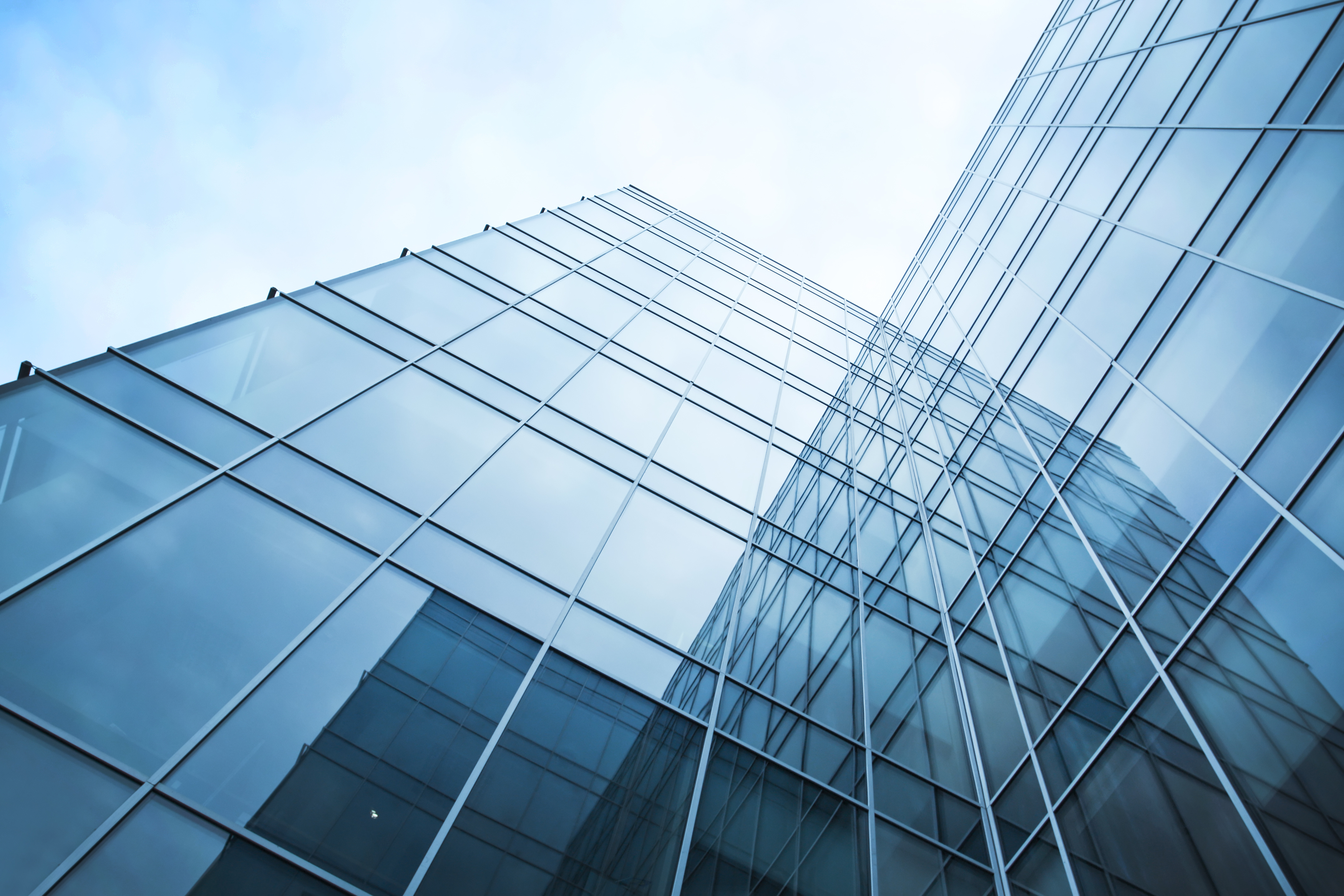 A modern glass building is shown from a low angle, highlighting its sleek, reflective exterior. The structure's vertical and diagonal lines create a dynamic sense of height against a blue sky, showcasing contemporary architectural design.