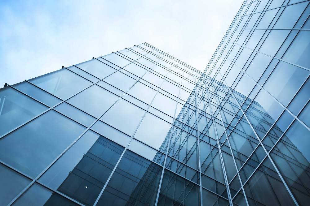 A modern glass skyscraper is viewed from below, showcasing its reflective surface and geometric lines against a cloudy sky. The building's design emphasizes transparency and symmetry, reaching upward into the atmosphere.