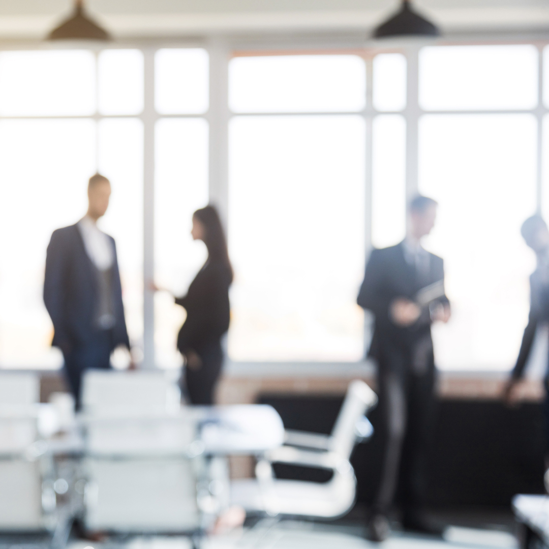 A group of blurred professionals in business attire engage in conversation within a bright office setting. Large windows provide natural light, creating an open and modern atmosphere. In the foreground, a conference table with empty chairs is partially visible.