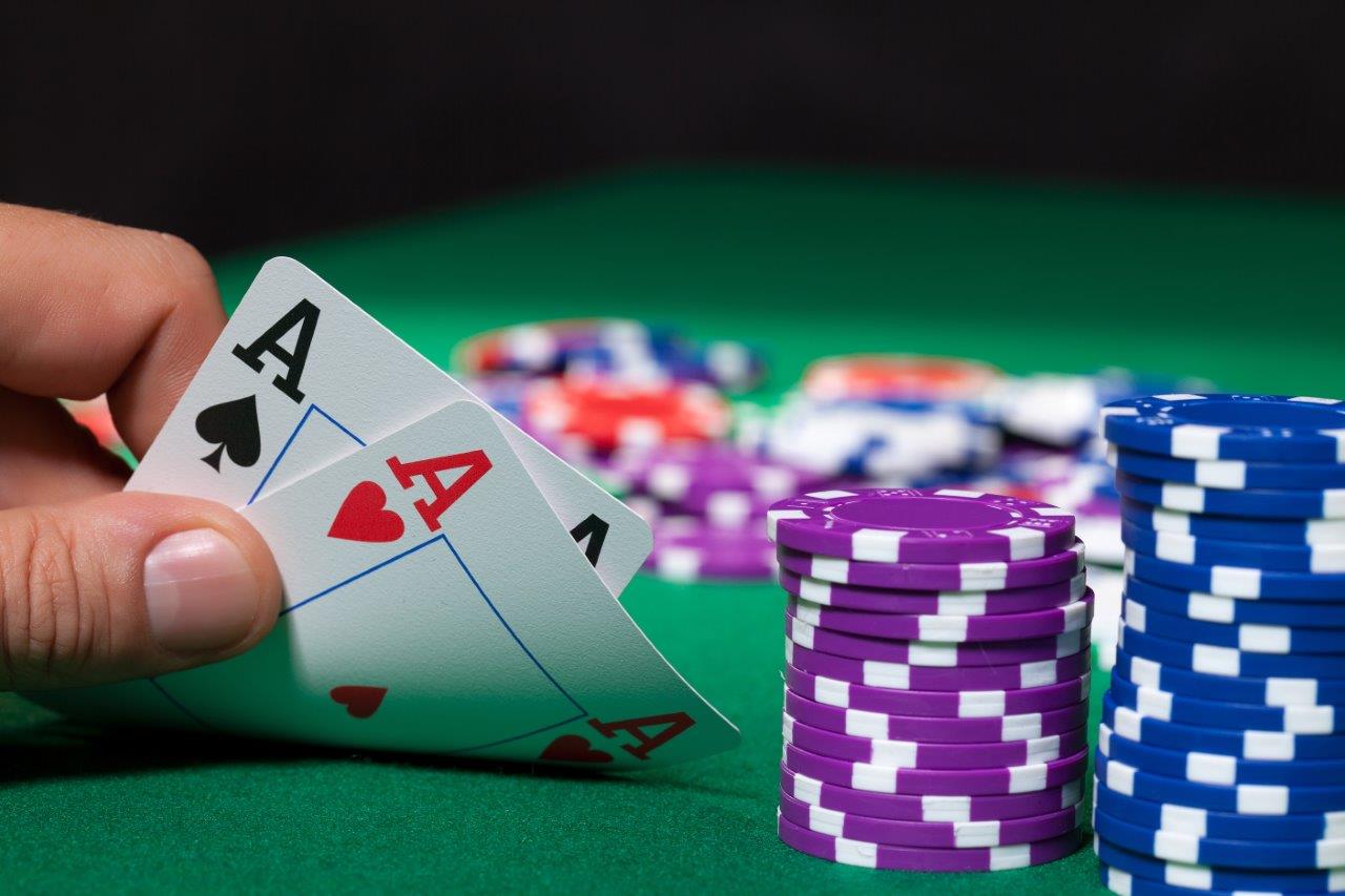 A hand is holding two playing cards, both showing an Ace—one in spades and the other in hearts. In the background, there are stacks of colorful poker chips on a green table.
