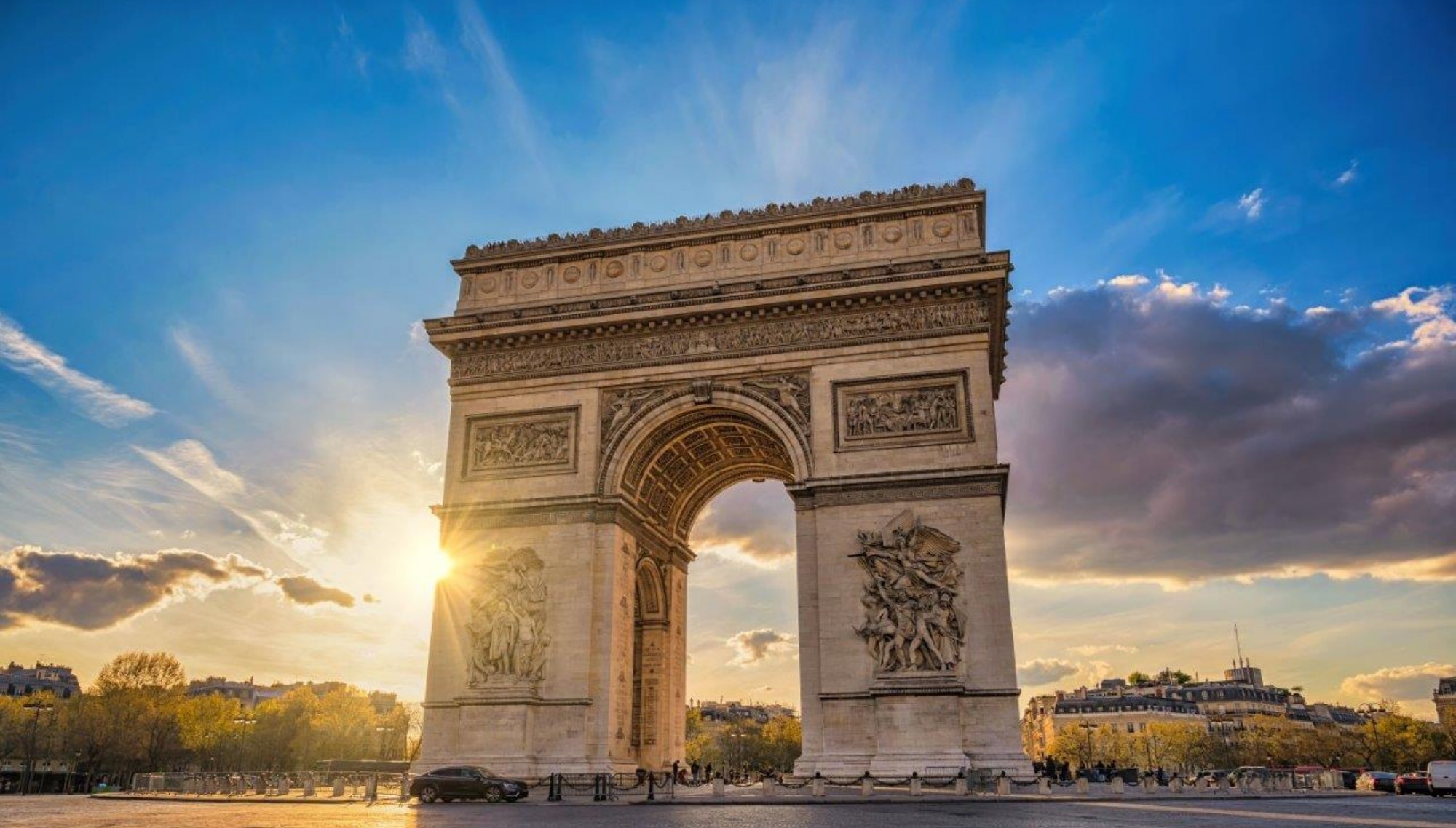 Der Bogen des Triumphs in Paris zeigt kunstvolle Reliefs und steht majestätisch im Sonnenlicht. Die Szene vermittelt eine beeindruckende Atmosphäre mit einem strahlenden Himmel und Wolken im Hintergrund.