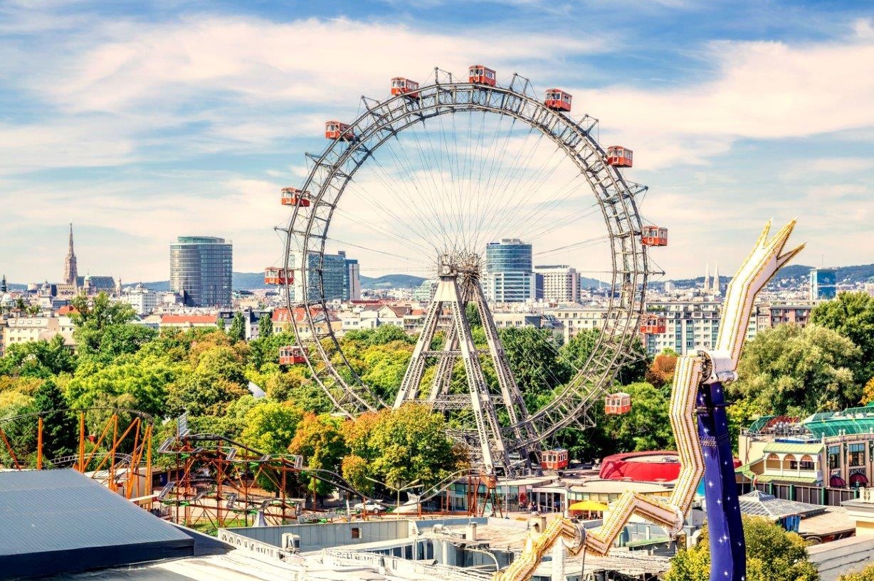 Ein Riesenrad mit roten Gondeln steht im Vordergrund, umgeben von einem Vergnügungspark und grünen Bäumen. Im Hintergrund sind moderne Gebäude und ein klarer Himmel sichtbar. Die Szene vermittelt eine fröhliche und einladende Atmosphäre.