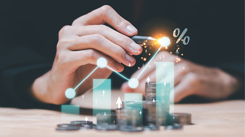 A hand holds a pen, tracing an upward trend on a graphic of bar charts and arrows, symbolizing financial growth. In the foreground are stacks of coins, reinforcing the theme of investment and increasing value.