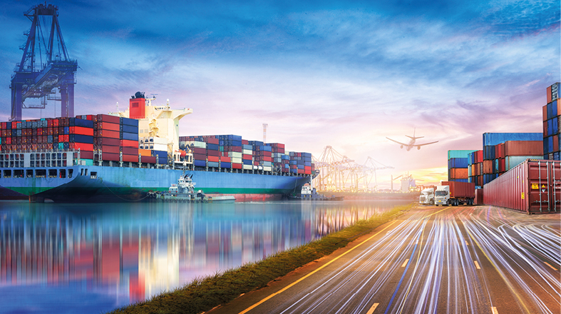 A large cargo ship docked at a bustling port, surrounded by stacked shipping containers. In the background, cranes are visible, and an airplane flies overhead. A road runs alongside the water, with lights suggesting traffic movement. The scene is set against a colorful sunset sky.