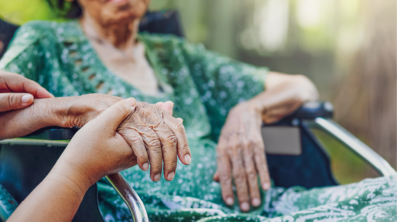 A close-up of two hands, one older and wrinkled, holding a younger hand gently. The background reveals a woman in a wheelchair, conveying a moment of affection and care between generations. The setting appears serene and supportive.
