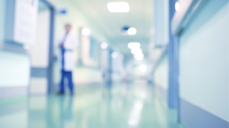 A blurred image of a hospital corridor shows a figure in a white coat standing near a doorway. The corridor has green flooring and is lined with other doorways, creating a clinical environment. Soft lighting illuminates the space.
