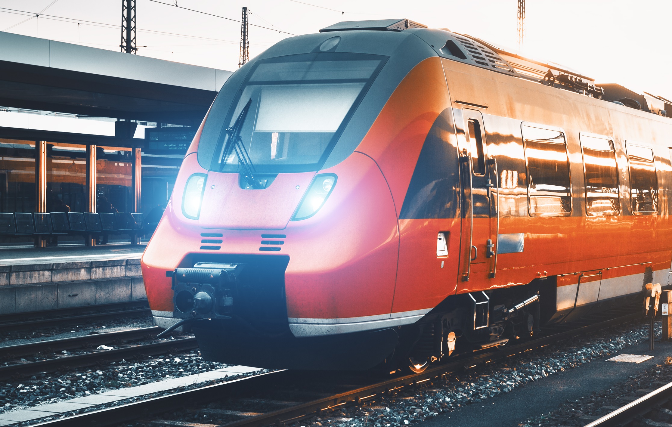 A red and black train is parked on the tracks at a station. Its front features bright headlights, and the train's sleek design reflects the light from the setting sun, highlighting its modern features. Empty benches are visible in the background.