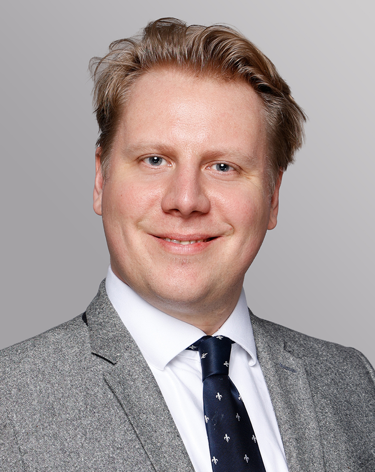 Porträt eines lächelnden Mannes mit blonden Haaren, der einen grauen Anzug und eine gestreifte Krawatte trägt. Der Hintergrund ist neutral und grau, was den Fokus auf das Gesicht des Mannes legt.