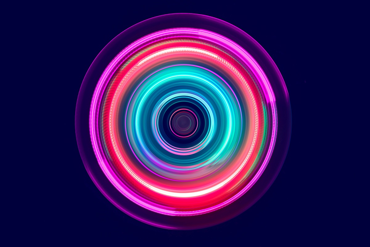 A vibrant, circular pattern featuring swirling colors of pink, blue, and white, giving a sense of movement and energy. The design creates an optical illusion of depth, with concentric rings radiating outward from the center against a dark background.