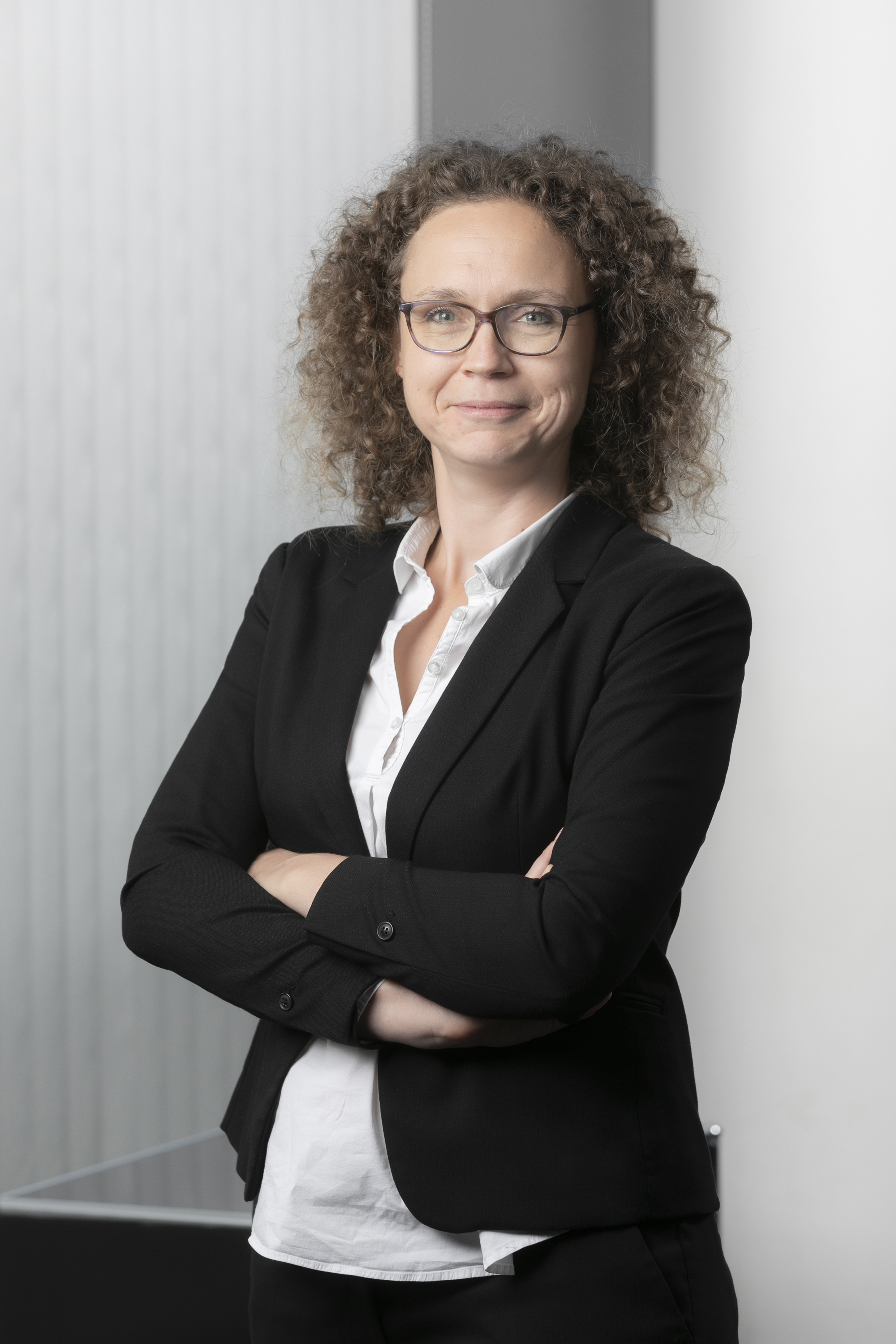 A professional woman with curly hair and glasses stands with her arms crossed, wearing a black blazer over a white shirt. She is smiling confidently against a minimalistic background, conveying a sense of competence and approachability.