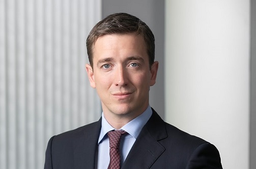 A professional-looking man in a suit and tie poses confidently. He has short brown hair and a slight smile, with a neutral background highlighting his formal appearance. The image conveys a sense of professionalism and approachability.