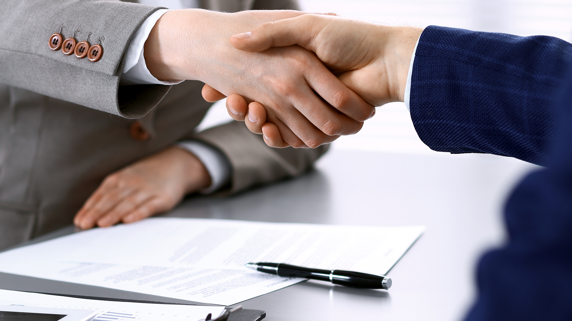 Two hands are shaking, symbolizing a formal agreement or partnership. In the background, there are documents and a black pen on a table. The setting suggests a professional or business environment.