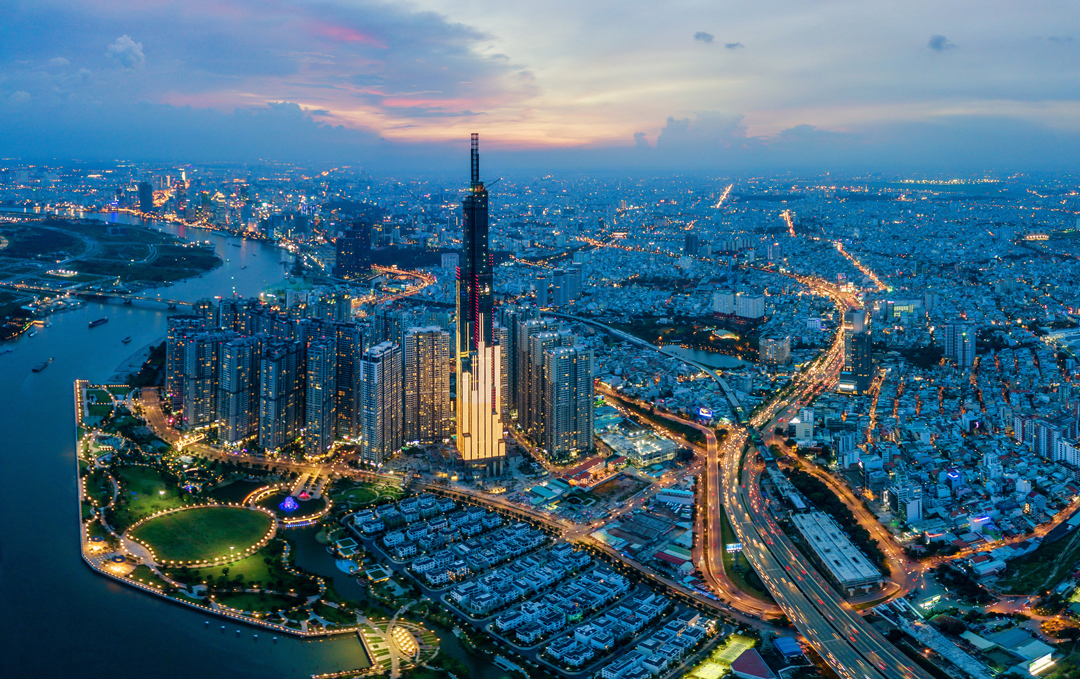 A panoramic view of a vibrant city skyline at dusk, featuring the tall Landmark 81 building illuminated against the evening sky. Surrounding structures and winding roads are visible, with lights twinkling throughout the area, reflecting the dynamic urban landscape.