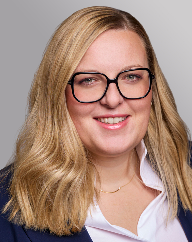 Porträt einer Frau mit langen, blonden Haaren und Brille, die in die Kamera lächelt. Sie trägt ein Business-Outfit in dunklem Blau mit einem weißen Hemd. Der Hintergrund ist neutral und grau.