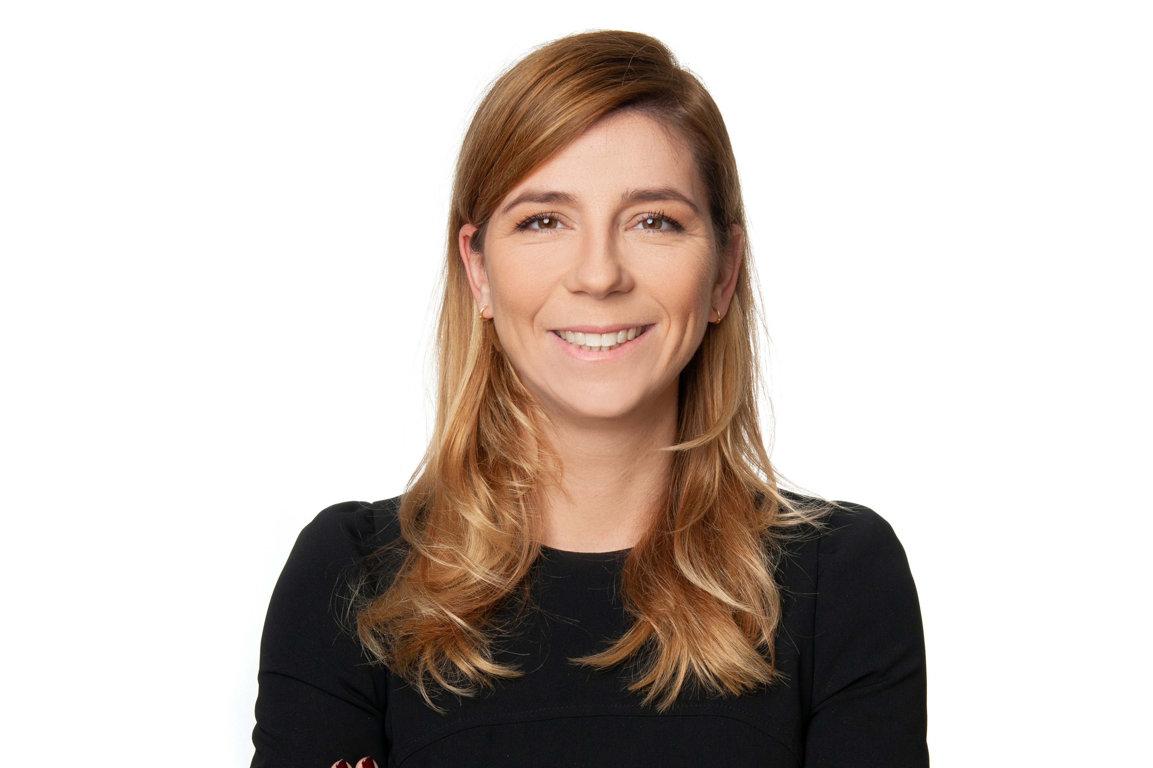 A smiling woman with long, wavy blonde hair is wearing a black top. She is standing with her arms crossed, against a plain white background. The overall expression is friendly and approachable.