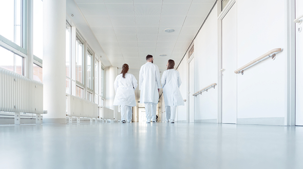 Three medical professionals in white lab coats walk down a bright, empty corridor. They are seen from behind, moving toward a set of doors at the end of the hallway, emphasizing a calm and professional environment in a healthcare setting.