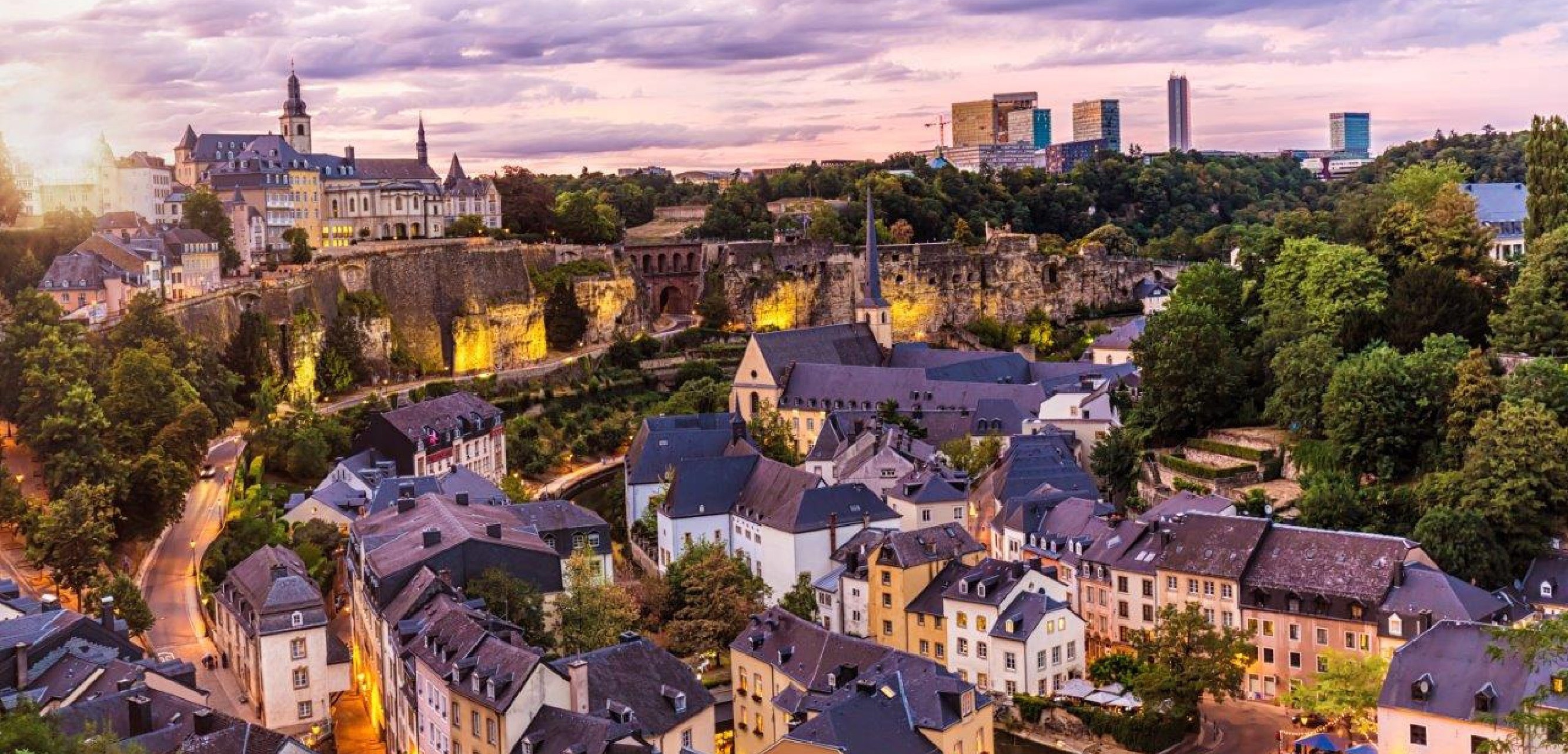 Eine Panoramaansicht von Luxemburg-Stadt bei Sonnenuntergang. Im Vordergrund sind Häuser mit traditionellen Dächern zu sehen, während im Hintergrund moderne Gebäude sowie eine Festung und eine Kirche hervorstechen. Die Landschaft wirkt grün und einladend, mit einem dramatischen Himmel.