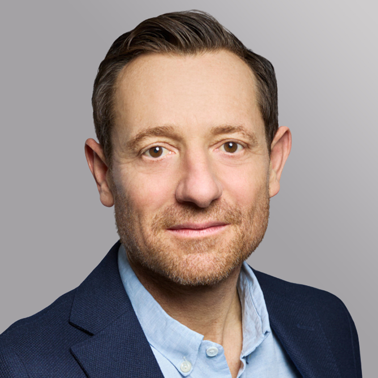 Porträt eines Mannes mit kurzem, hellbraunem Haar, einer leichten Stoppelbart und einem neutralen Ausdruck. Er trägt einen dunkelblauen Blazer und ein hellblaues Hemd. Der Hintergrund ist schlicht und einfarbig.