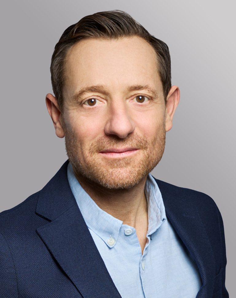 Ein Mann mit kurzen, braunen Haaren und einem Bart schaut in die Kamera. Er trägt einen blauen Blazer und ein hellblaues Hemd. Der Hintergrund ist grau und einfarbig.