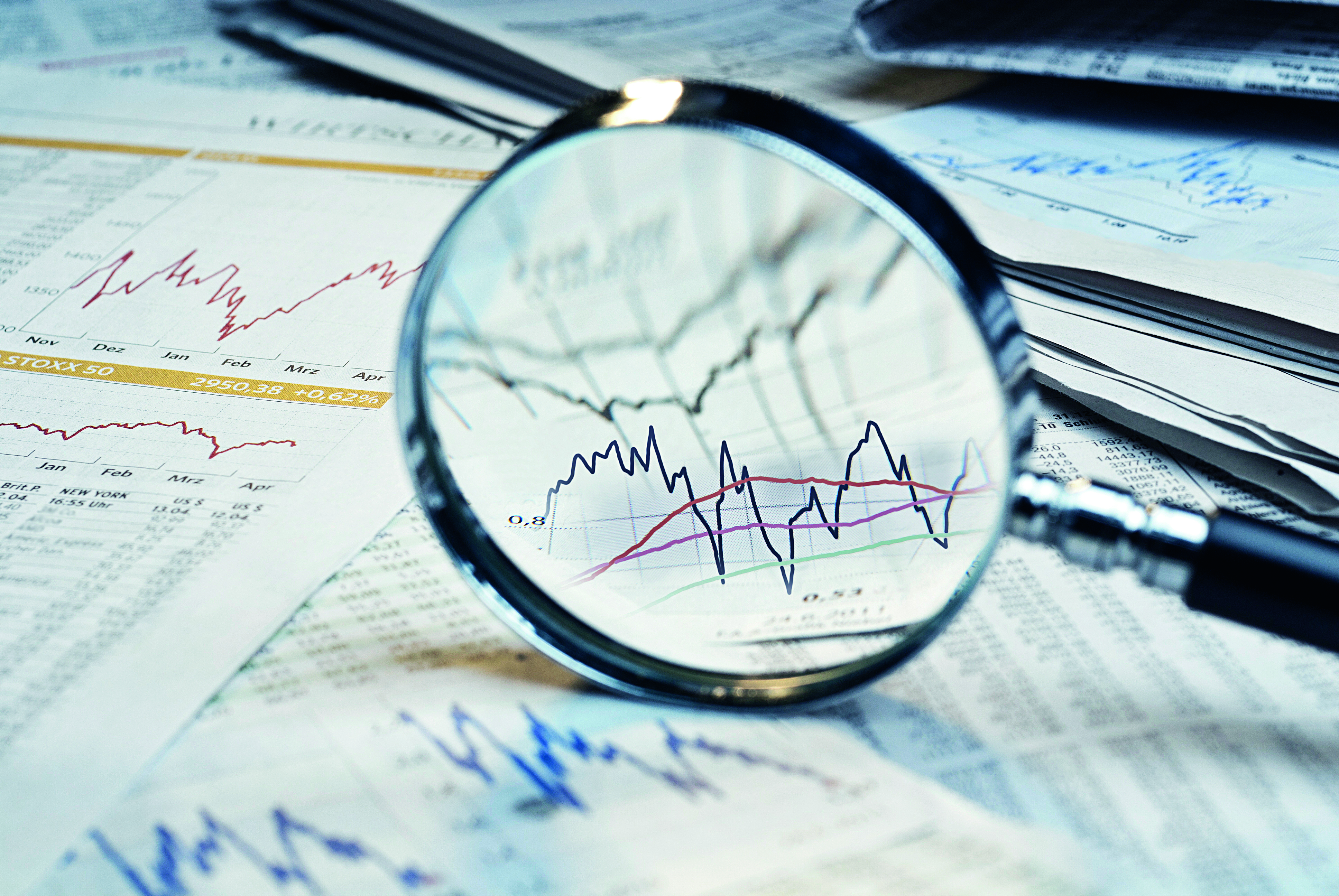 A magnifying glass focuses on stock market graphs and charts scattered on a table. The graphs show fluctuating lines representing financial data, emphasizing analysis and market trends. The surrounding papers feature various financial statistics and data points, highlighting a financial research context.